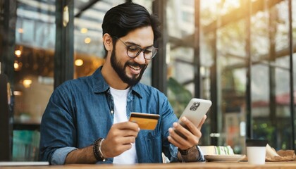 Bearded handsome man makes purchase online. A hipster wearing casual shirt holds a mobile phone and a credit card orders food. Smiling guy is using smartphone and debit bank card for paying in e-shop