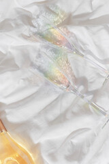 Rainbow color shining champagne glasses and white sparkling wine bottle on bed, on white blanket background. Lifestyle aesthetic photo, star filter. Romance meeting, romantic holiday concept.