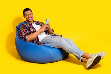 Full length photo of relaxed guy wear checkered shirt lay on bean bag hold smartphone in hands isolated on vibrant color background