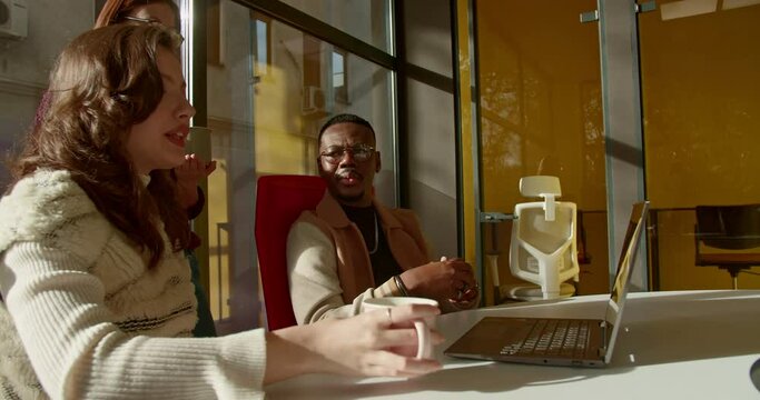 A group of colleagues discussing project ideas in a bright, modern office space, sipping coffee and using a laptop for research.