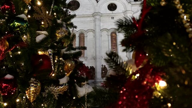 Close-up view of colourful Christmas baubles and light garland hanging on Christmas trees next to Cathedral of Christ the Saviour in a dark winter night in Moscow, Russia. Slow motion handheld video.