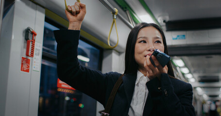 Pretty Asian woman in formal suit using mobile phone while standing in tram or bus. Young female...