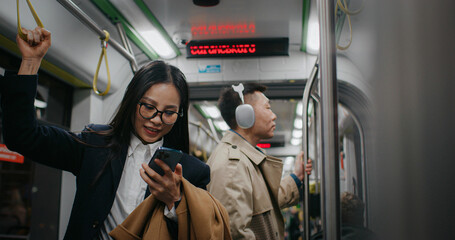 Serious Asian businesswoman holding on to safety handle with one arm. People in background standing still and listening to music in headphones in public transport.