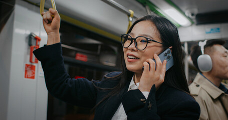 Beautiful Chinese woman standing in middle of train. Talking on mobile phone with someone. Agreeing for next meeting or creating plans for weekend. Wearing official attire and large glasses.