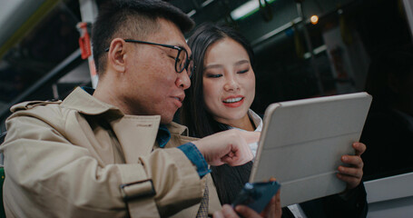 Charming Chinese couple sitting next to each other in moving bus or train. Girl getting attention from man by tapping shoulder. Woman showing information on screen of tablet to husband or boyfriend.