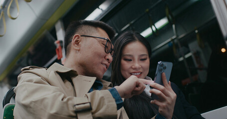 Friends sitting close inside moving bus or train at night. Evening trip. Going back home from work. Watching videos on smartphone. Man pointing on phone screen for friend. Discussing video or image.