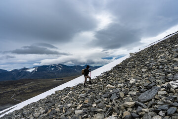 Male traveler in the snowy mountains of Norway. Young sporty Man climbing on top during trekking hiking with Scandinavian sticks. Hike in stony rocks mountainous area. Amazing northern cloudy view.