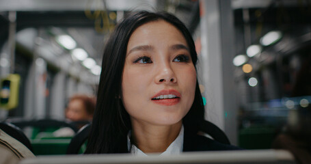 Beautiful Asian woman sitting next to window in bus or train. Girl with straight black hair wearing many earrings. Checking outside to see stop name. Continuing having fun with digital device.