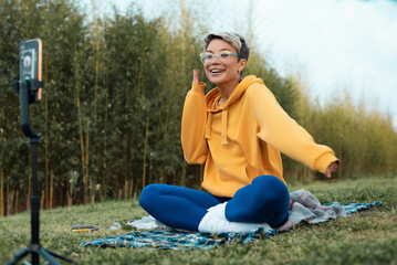 a pretty blogger girl in glasses and an orange hoodie is sitting in a park in nature, streaming on...