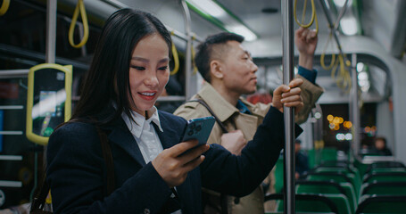 Chinese young girl standing in train or long bus. Using smartphone in public transport. Laughing from video or text. Typing something on sensor with one finger. Holding balance with left hand.