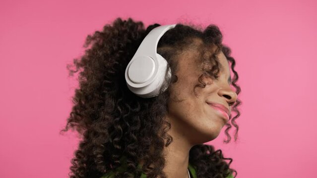 Happy mixed race woman listening music, enjoying headphones, pink background