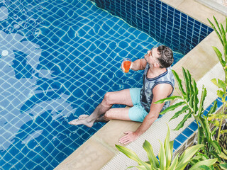 Handsome man with a glass of drink sits near the swimming pool. Sunny, clear morning. View from above. Closeup, outdoors. Vacation and travel concept