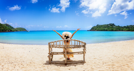 A woman with sunhat sits on a driftwood bench at the tropical Carlisle Bay beach, Antigua and...
