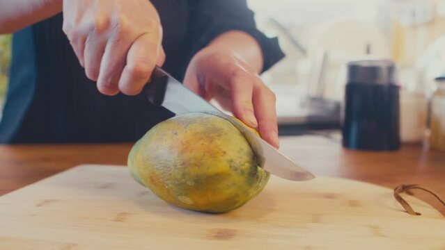 Close up of woman's hands cutting papaya fruit, pawpaw fruit in half using a knife. Female cook preparing papaya, chef in a brightly lit kitchen. Studio shot. High quality 4k footage