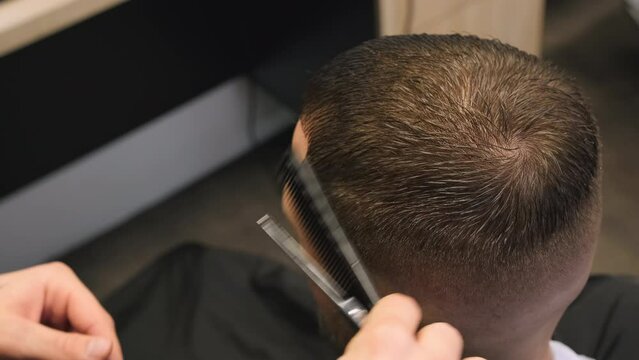Barber cuts client wet hair with comb holding scissors in barbershop closeup. Master serves professional haircare to man in beauty salon