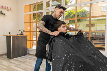 peluquero,barbero tatuado  cortando el pelo , la barba y el bigote a un cliente joven y moreno