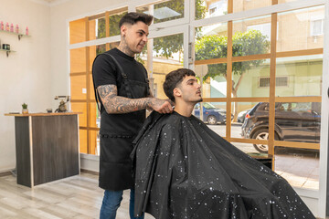 peluquero,barbero tatuado  cortando el pelo , la barba y el bigote a un cliente joven y moreno