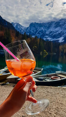 Person holding cocktail with view of colorful wooden boats on Fusine Lake (Laghi di Fusine) in...