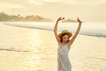 Woman in polka dot dress enjoys sunset at beach, arms raised high. Solo traveler celebrates...
