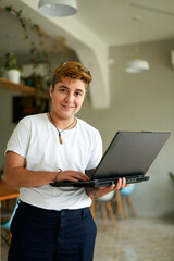 Confident transgender employee holds laptop in modern office cafe. Short-haired, smiling FTM pro in...