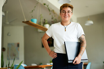 Transgender professional in bright office smiles, holding laptop, inked arms visible. Confident stance inclusive work culture advocate. Nerdy glasses smart casual work attire. Plants modern workspace.