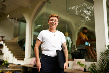 Smiling transgender pro holds laptop in modern workspace. Inclusive work environment with diverse...