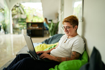 Transgender person lounges on bean bag chair, working on laptop in a light-filled coworking space, reflecting modern work flexibility and inclusion in the professional environment.