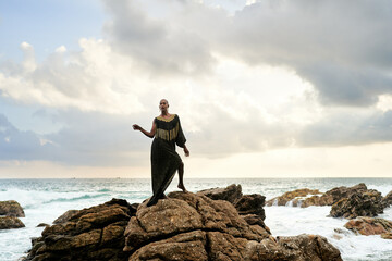 Gender fluid black person poses gracefully standing on rocks in ocean . Androgynous ethnic fashion model in posh dress and jewellery on rocky beach by storm. Pride month
