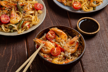 Rice with shrimp and udon in Pad Thai sauce on a wooden table next to cherry tomatoes, soy sauce and chopsticks