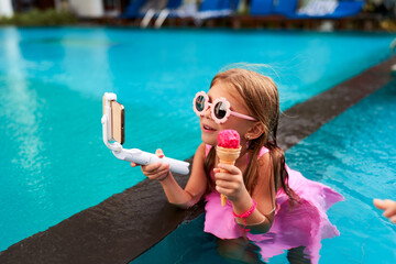 Little girl enjoys ice cream, streams live in pool. Wearing pink swimwear, heart-shaped sunglasses,...