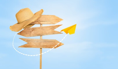 wooden road sign with a beach hat on the edge and a flying paper airplane against the sky. travel...
