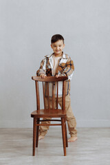 A young boy is standing in front of a wooden chair. He is wearing a plaid jacket and tan pants. The...