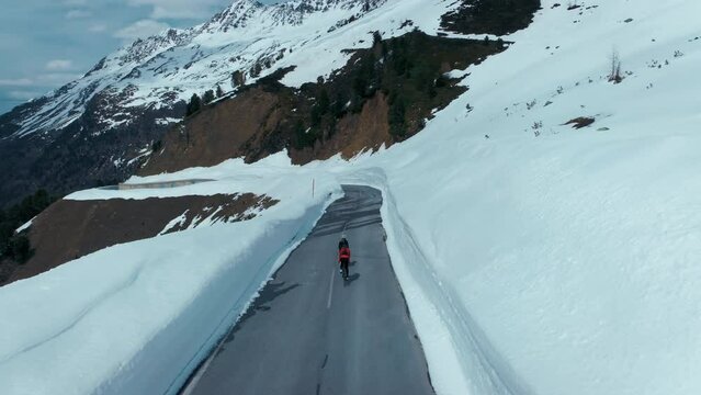 Two cyclists descend epic mountain road on sunny spring day, ride in snow walls corridor into the valley. Winter and cold conditions cycling