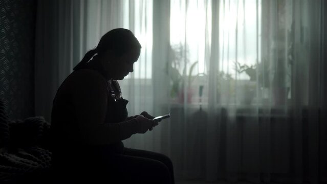 Silhouette of isolated woman scrolling on smartphone in dim room by window. Alone female engages with tech, lost in virtual world, disconnected from reality. Indoor solitude captured in shadows.