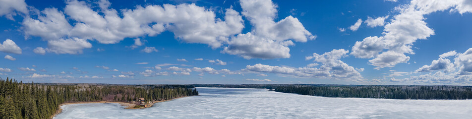 Panoramic view of a frozen lake that is surrounded by spruce trees. The sky is bright blue with puffy white spring clouds. A cabin can be seen on the shoreline.
