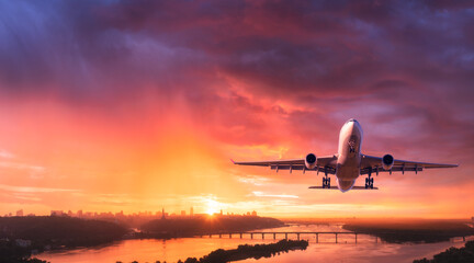 Fototapeta na wymiar Airplane is flying in colorful sky over city at sunset. Landscape with passenger airplane, skyline, purple sky with red and pink clouds at dusk. Aircraft is landing at twilight. Aerial view of plane