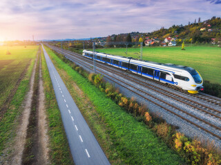 Aerial view of modern high speed train moving near alpine mountains and green fields, bike path at sunset in spring. Top view of train, railroad, road, meadows in summer. Railway station in Croatia