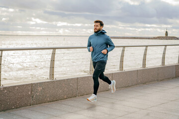 A bearded man in a teal jacket finds his pace along a waterfront promenade, clouds gathering above.