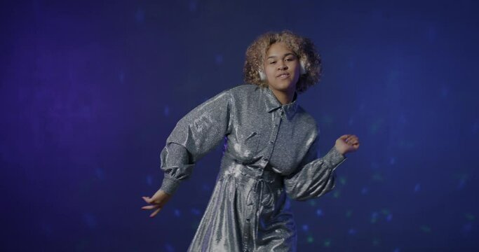 Slow motion portrait of African American performer wearing headphones dancing enjoying music on blue background. Woman wearing trendy outfit moving to pop song.