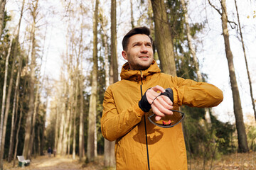 A runner in a golden jacket pauses to check his watch on a sunlit forest path, breath visible in...