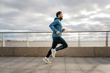A determined athlete runs on a waterfront promenade, clouds above hinting at imminent weather.
