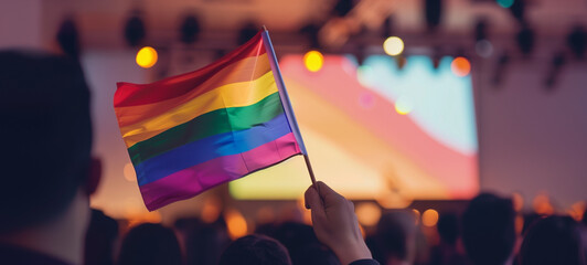 A person waving a rainbow flag at an event, with a big screen in the background