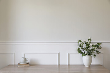 Ceramic vase with blooming apple tree branches. Cup of coffee, tea on wooden table, desk with old books. Scandi home interior. Spring breakfast still life. Empty white wall mockup, stucco decor.