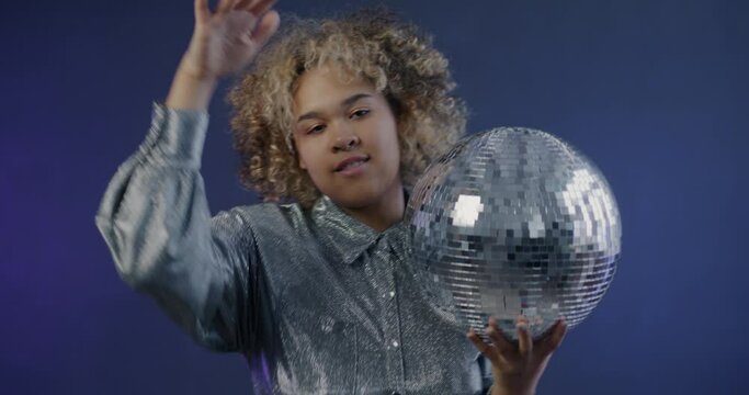 Slow motion portrait of excited African American girl dancing with disco ball having fun at party on blue background. Entertainment and millennial concept.