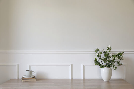 Ceramic vase with blooming apple tree branches. Cup of coffee, tea on wooden table, desk with old books. Scandi home interior. Spring breakfast still life. Empty white wall mockup, stucco decor.