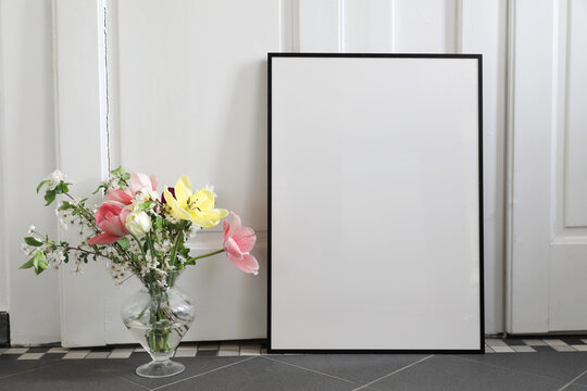 Spring still life scene. Tulips, cherry tree blossoms bouquet in glass vase. Blank black picture frame, poster mockup on grey tile floor. White wall background . Artisitic Easter decor, interior home.