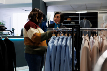 Clothing store woman asian assistant talking and consulting customer. Customer exploring rack with...