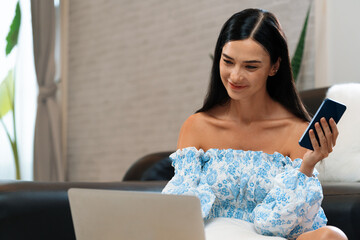 Young woman using laptop, happily browsing the internet or making online purchases on sofa at home....