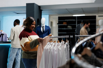 Smiling african american woman holding jacket on hanger while shopping for outfit in clothing...