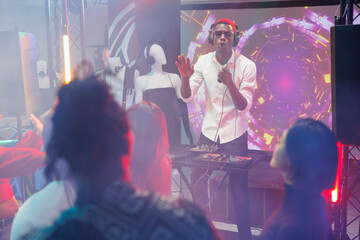 Dj playing on stage while people having fun at disco party in nightclub. Young african american man performing electronic music for clubbers crowd at discotheque in club with spotlights